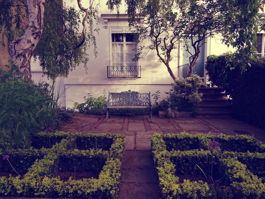 A small urban garden featuring a paved patio area with a decorative metal bench positioned against a white house wall. The garden is bordered by well-maintained planting beds with neatly trimmed boxwo