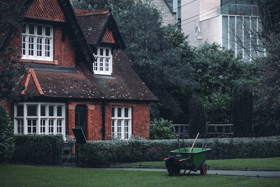 A small urban garden featuring a paved patio area with a decorative metal bench positioned against a white house wall. The garden is bordered by well-maintained planting beds with neatly trimmed boxwo