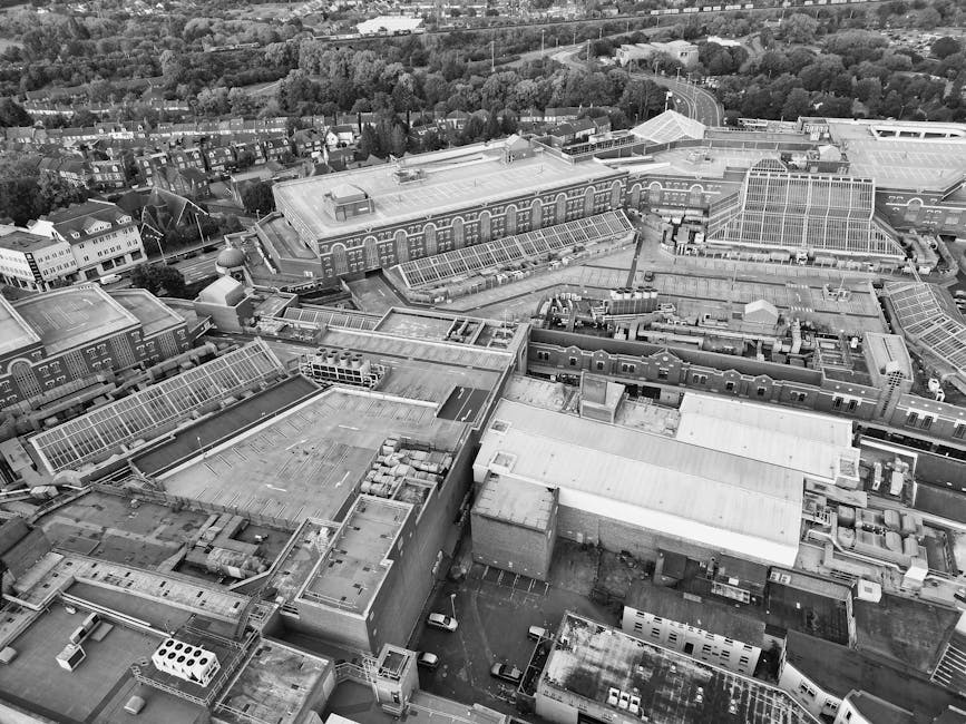 An aerial black-and-white photograph of a large urban shopping centre, featuring multiple interconnected flat-roofed buildings with glass roof sections, solar panels, and ventilation units. Surroundin
